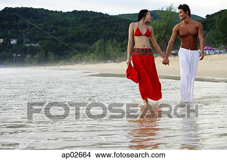 Picture - Couple walking side by side along beach, holding hands, looking at each other . Fotosearch
