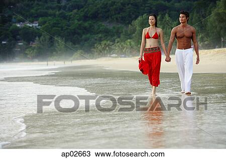 Stock Image - Couple walking side by side along beach, holding hands . Fotosearch