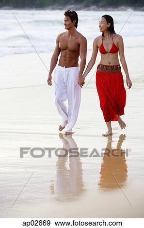Stock Photo - Couple walking side by side along beach, holding hands, looking away . Fotosearch