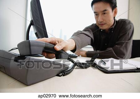 Stock Image - Male executive sitting at office desk, reaching for telephone . Fotosearch