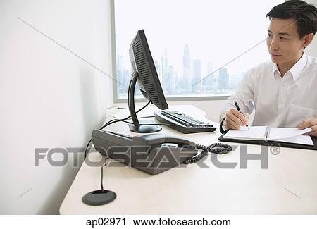 Stock Image - Male executive sitting at office desk, writing in organizer, looking away. Fotosearch