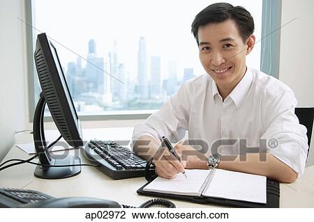 Stock Image - Male executive sitting at office desk, looking at camera, smiling. Fotosearch