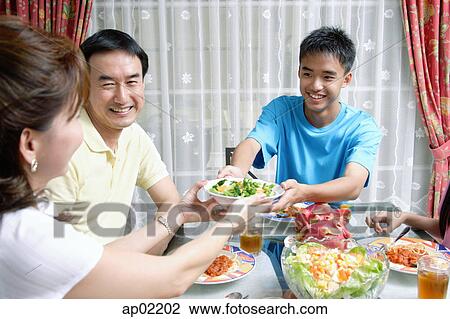 Stock Image - Mother and son passing food to each other at dining table . Fotosearch