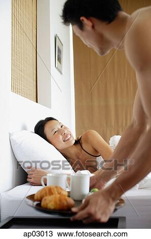 Stock Image - Woman in bed, man setting down breakfast tray on bedside table . Fotosearch