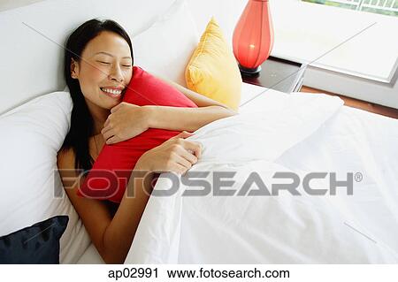 Stock Image - Woman lying in bed, hugging pillow, eyes closed . Fotosearch