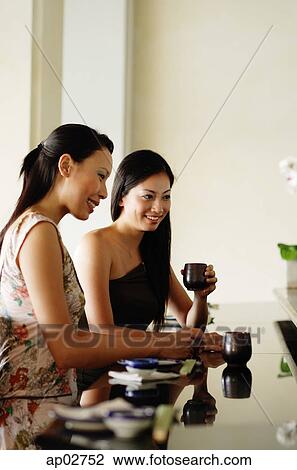 Stock Image - Women sitting at table, side by side, having Chinese tea . Fotosearch