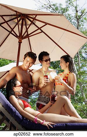 Stock Image - Young adults standing and holding drinks, one woman sitting on deck chair . Fotosearch