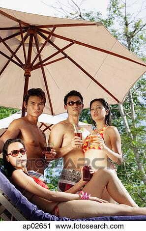 Young adults standing under umbrella, holding drinks, one woman sitting on deck chair View Large Photo Image Stock Image - Young adults standing under umbrella, holding drinks, one woman sitting on deck chair . Fotosearch