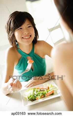 Stock Image - Young woman at table, smiling at person in front of her . Fotosearch