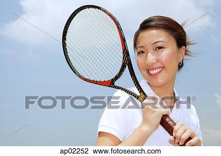 Stock Image - Young woman holding tennis racket, smiling at camera . Fotosearch