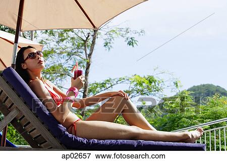 Stock Photo - Young woman in bikini, lying on deck chair, holding drink . Fotosearch