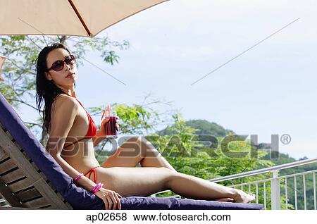Stock Photo - Young woman in bikini, lying on deck chair, holding drink, turning to look over shoulder . Fotosearch