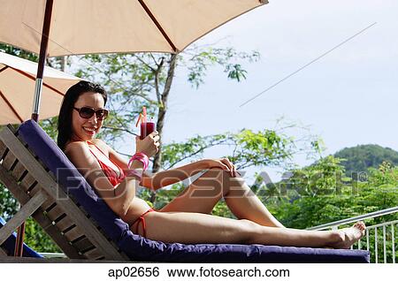 Young woman in bikini, lying on deck chair, holding drink, smiling at camera View Large Photo Image Stock Photograph - Young woman in bikini, lying on deck chair, holding drink, smiling at camera. Fotosearch