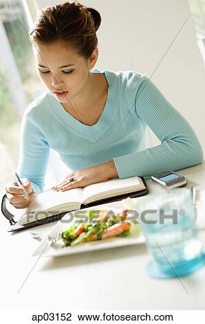 Young woman sitting at table, food next to her, writing in notebook View Large Photo Image Stock Image - Young woman sitting at table, food next to her, writing in notebook . Fotosearch