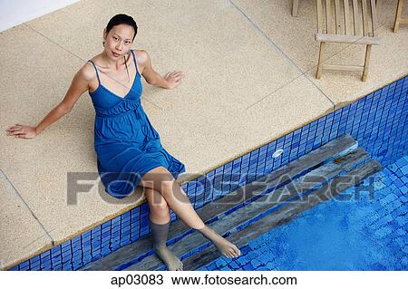 Stock Image - Young woman sitting by swimming pool, feet in water, looking at camera . Fotosearch