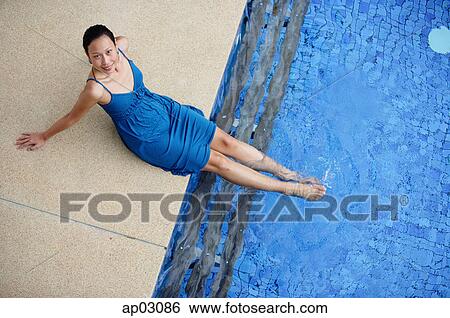 Stock Photograph - Young woman sitting by swimming pool, feet in water, smiling at camera . Fotosearch
