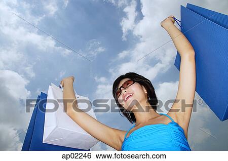 Stock Photograph - Young woman with arms outstretched holding shopping bags, low angle view . Fotosearch
