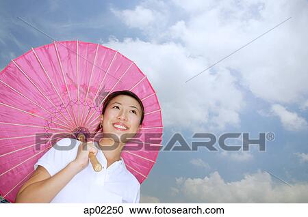 Stock Image - Young woman with pink umbrella, smiling, low angle view . Fotosearch