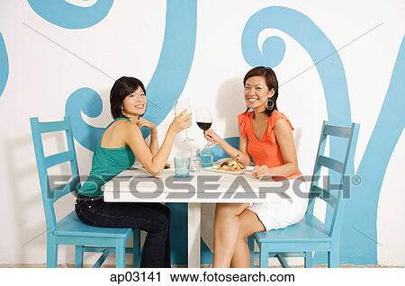 Stock Image - Young women in cafe, toasting with wine glasses, smiling at camera . Fotosearch