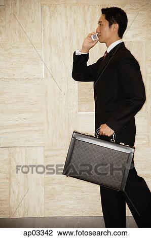 Stock Image - Businessman carrying briefcase, using mobile phone. Fotosearch