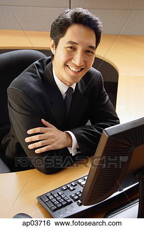 Stock Photograph - Businessman sitting at desk, arms crossed, smiling at camera . Fotosearch