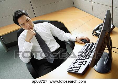 Businessman sitting at desk, looking at computer View Large Photo Image Stock Photo - Businessman sitting at desk, looking at computer . Fotosearch