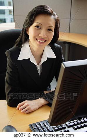 Stock Image - Businesswoman sitting at desk, smiling at camera . Fotosearch