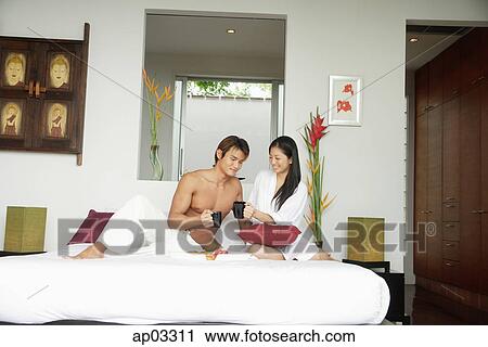Stock Image - Couple in bedroom, sitting on bed, holding mugs. Fotosearch