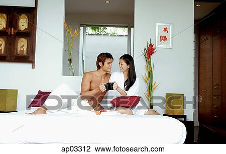 Stock Image - Couple in bedroom, sitting on bed, toasting with mugs . Fotosearch