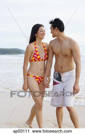 Stock Image - Couple on beach, holding hands, looking at each other. Fotosearch
