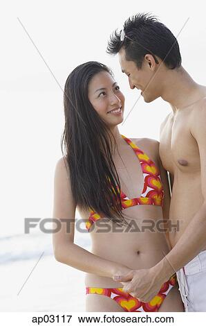 Stock Photo - Couple on beach, holding hands looking at each other. Fotosearch