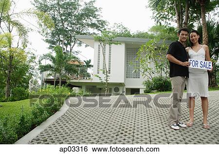 Stock Photograph - Couple standing on driveway holding for sale sign. Fotosearch