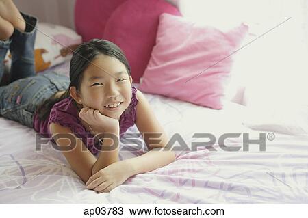Stock Image - Girl lying on bed, hands on chin, smiling at camera . Fotosearch