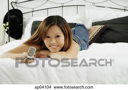 Picture - Girl lying on bed, holding mobile phone, looking at camera . Fotosearch