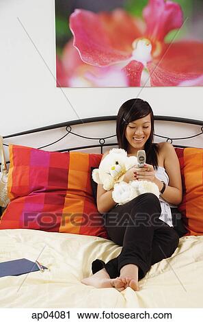 Stock Image - Girl sitting on bed, hugging stuffed toy, using mobile phone. Fotosearch