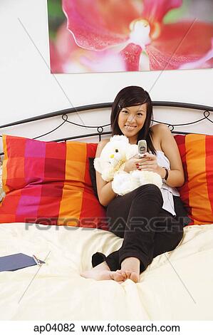 Stock Image - Girl sitting on bed, hugging stuffed toy, using mobile phone, smiling at camera . Fotosearch