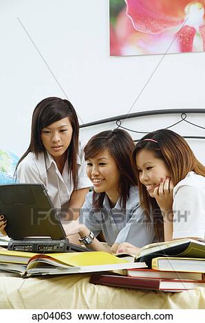 Stock Image - Girls in school uniform sitting side by side on bed, reading books . Fotosearch