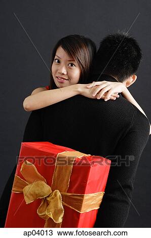 Stock Image - Man holding gift behind his back, woman embracing him, looking over shoulder at camera . Fotosearch
