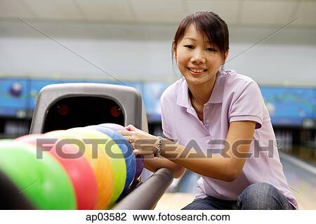 Stock Image - Woman selecting ball at bowling alley, smiling at camera . Fotosearch