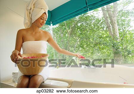 Stock Image - Woman sitting at edge of bath tub, throwing flower petals into tub . Fotosearch