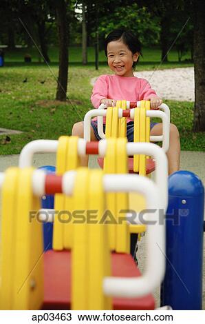 Stock Image - Young girl sitting on seesaw, smiling at camera . Fotosearch
