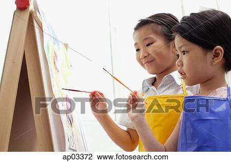 Stock Image - Young girls standing side by side, painting on easel . Fotosearch