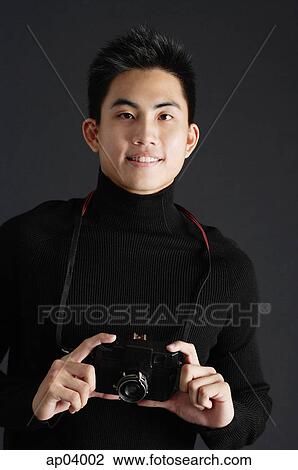 Young man dressed in black, standing against black background, holding camera View Large Photo Image Stock Image - Young man dressed in black, standing against black background, holding camera . Fotosearch