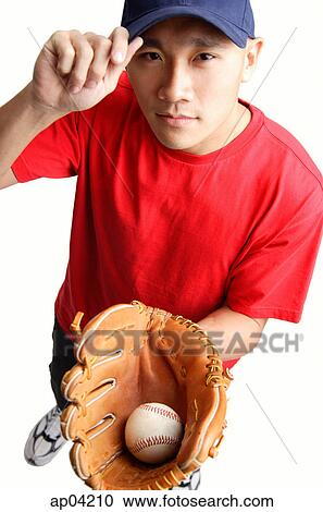 Stock Image - Young man holding baseball glove and ball, adjusting cap. Fotosearch