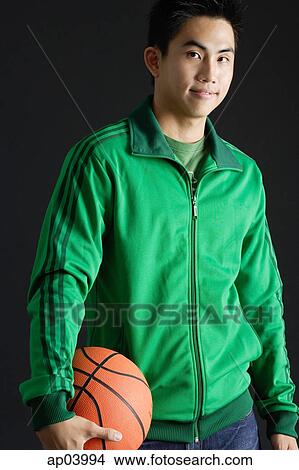 Young man wearing green tracksuit jacket, holding basketball, looking at camera View Large Photo Image Picture - Young man wearing green tracksuit jacket, holding basketball, looking at camera . Fotosearch