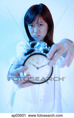 Young woman holding alarm clock, looking at camera, frowning View Large Photo Image Stock Image - Young woman holding alarm clock, looking at camera, frowning. Fotosearch
