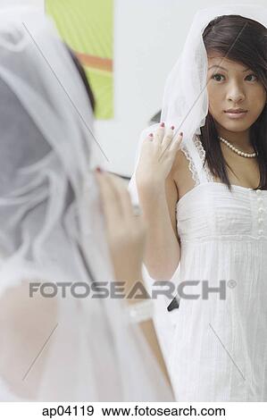 Stock Photo - Young woman in white dress and veil, looking at herself in mirror . Fotosearch