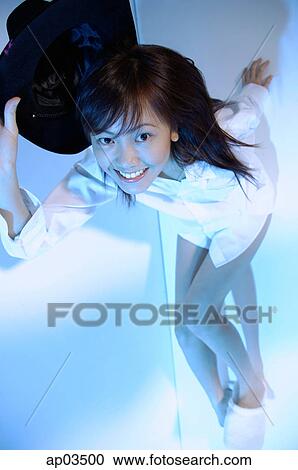 Stock Image - Young woman sitting on floor, looking at camera, holding hat. Fotosearch
