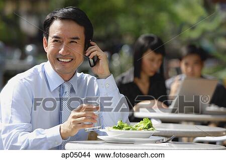 Picture - Businessman having lunch at outdoor cafe, using mobile phone, smiling. Fotosearch