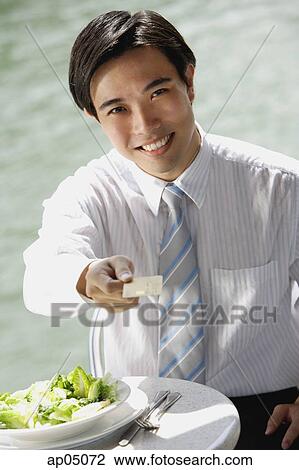 Businessman sitting at outdoor cafe, holding credit card towards camera View Large Photo Image Stock Image - Businessman sitting at outdoor cafe, holding credit card towards camera. Fotosearch